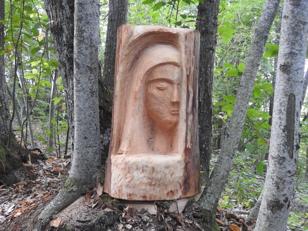 La figura emerge da una nicchia scavata nel tronco, che ne incornicia il volto come un arco naturale. Il viso, dai lineamenti aggraziati e i capelli lunghi, &egrave; colto solo per met&agrave; dalla superficie di legno. L'unico occhio visibile &egrave; chiuso, in un'espressione di raccoglimento simile alla preghiera. Tutt'intorno, la corteccia e la superficie grezza del legno restano a vista, in contrasto con la levigatezza del volto scolpito