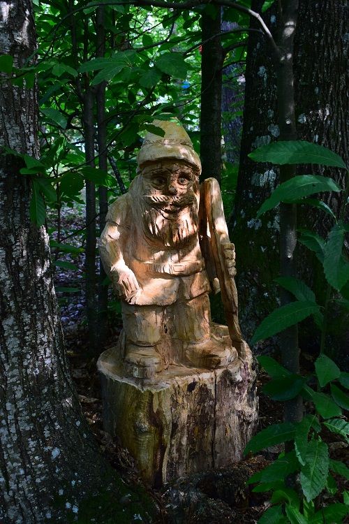 Piccola figura tozza di un folletto o gnomo, nascosta tra le piante del sottobosco. Indossa un cappello a punta e sfoggia una barba folta e lunga, con vestiti semplici. Il volto &egrave; scolpito con tratti semplici ma profondi. Con la mano sinistra la figura si appoggia a un lungo bastone.